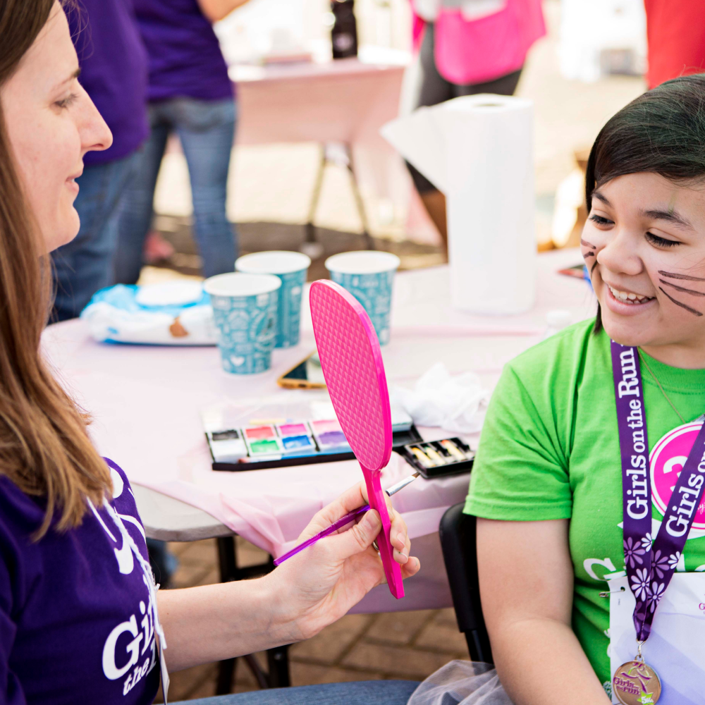A Girls on the Run volunteer holds up a mirror to a participant who has face paint on